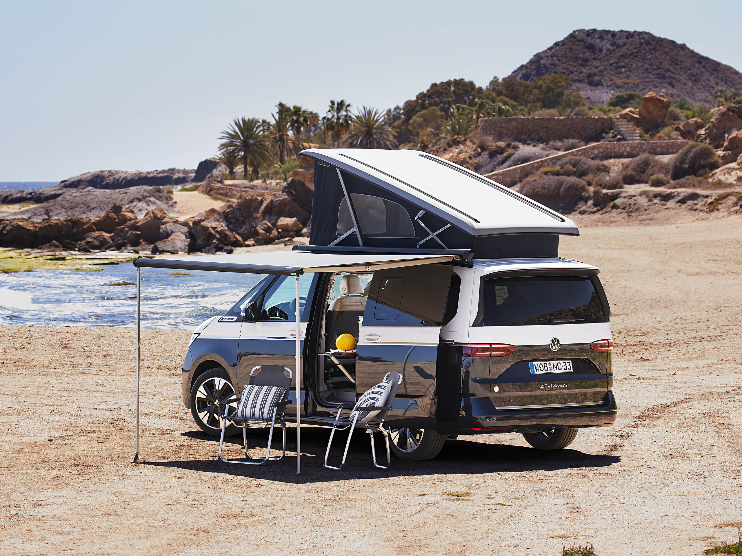 Een VW California met het uitklapbare dak omhoog en de luifel uitgeschoven en klapstoelen die op het strand staan.